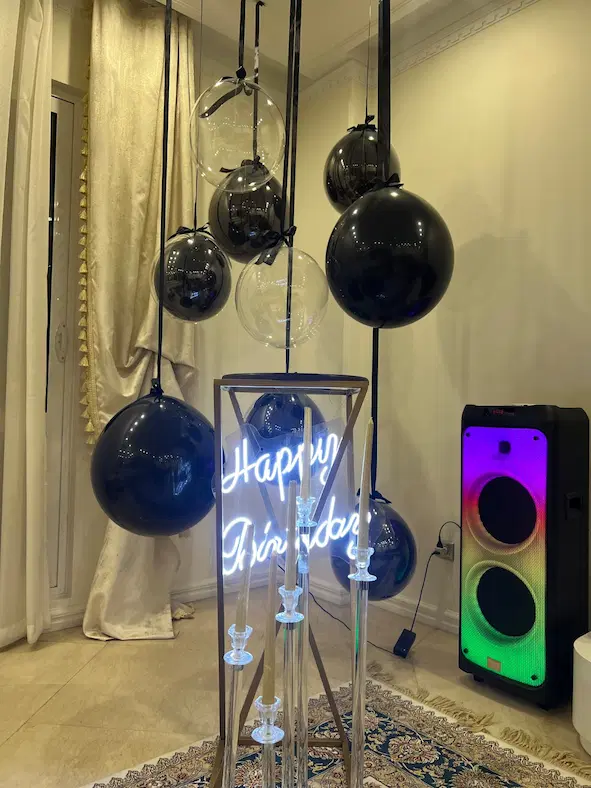 دکوراسیون مدرن تولد با بادکنکهای مشکی و شفاف آویزان، نئون Happy Birthday و شمعدانهای اکریلیک در فضای داخلی لوکس"
