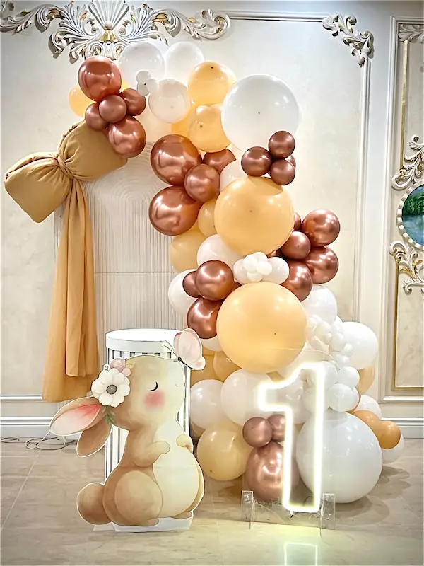 alt="دکوراسیون تم تولد خرگوش یکسالگی با بادکنکهای هلویی و مسی، نئون عدد ۱ و استند خرگوش فانتزی"