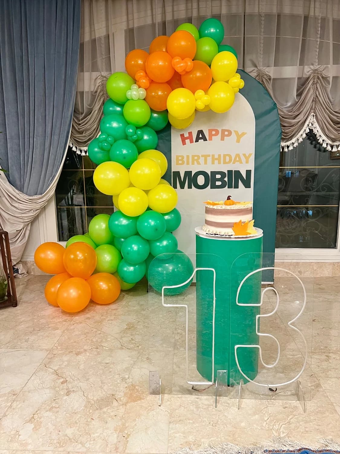 دکور تولد ۱۳ سالگی مبین با تم سبز و نارنجی شامل بکگراند سبز با نوشته Happy Birthday Mobin، آرک بادکنکی سبز زرد و نارنجی، پایه سبز با کیک و عدد اکریلیک ۱۳
