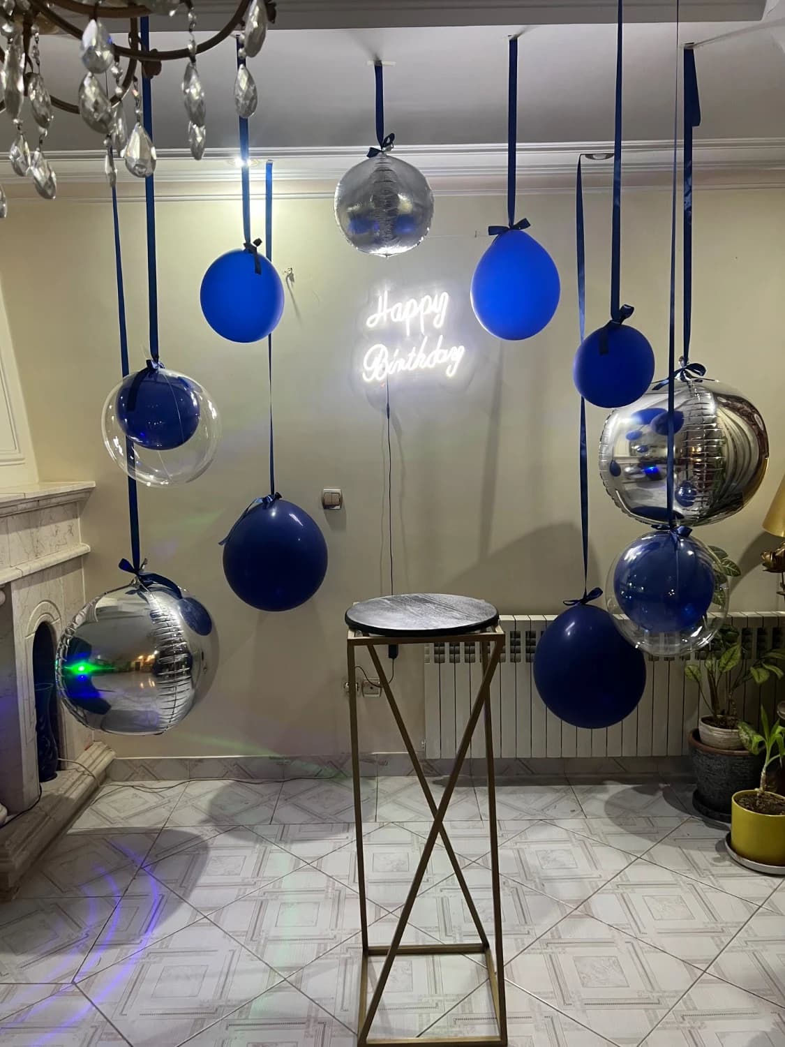 دکور تولد شیک آبی و نقرهای با نوشته نئونی Happy Birthday، بادکنکهای معلق آبی و کروم نقرهای با روبان آبی و استند طلایی در فضای داخلی