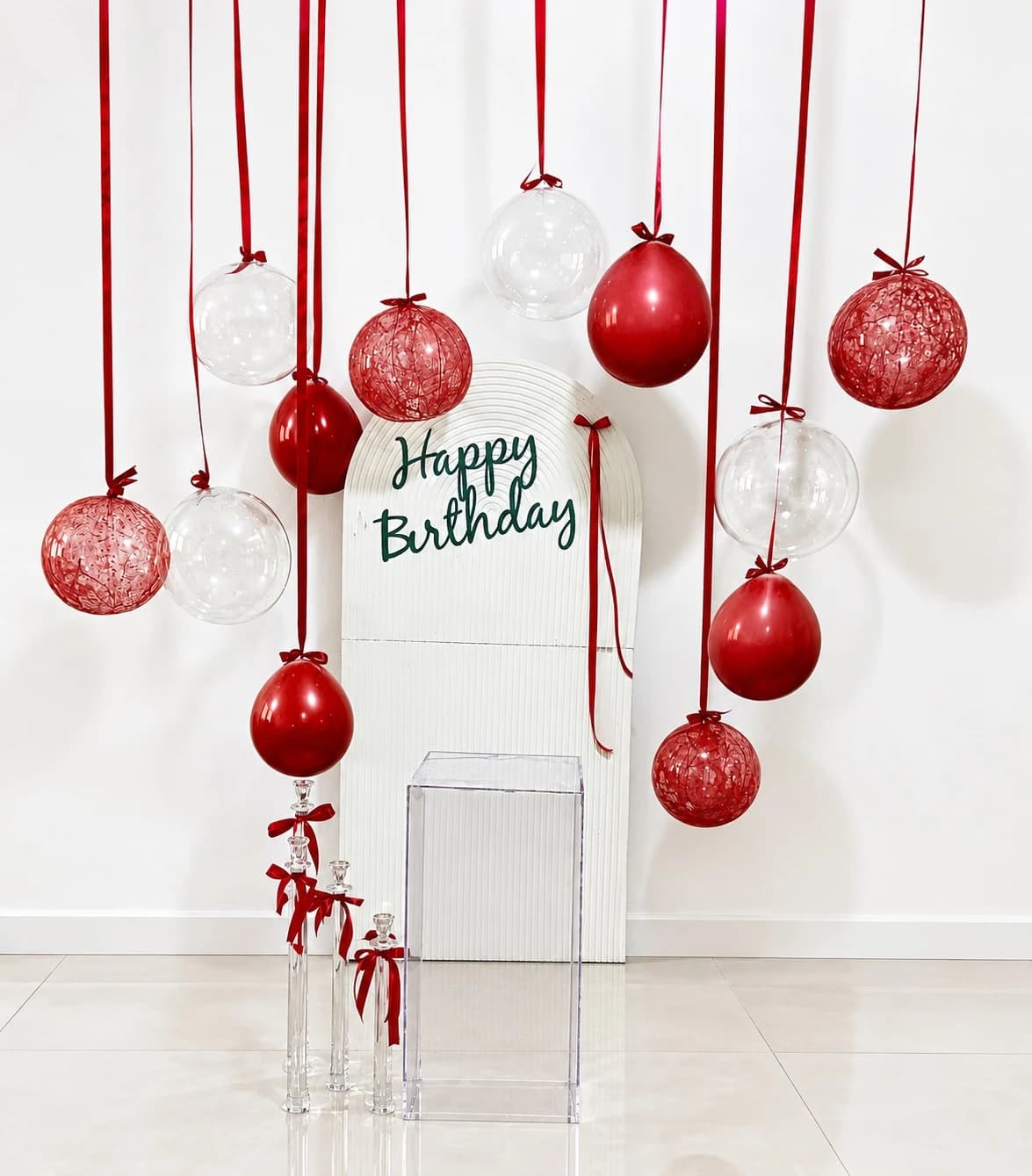 دکور تولد قرمز و سفید شیک شامل بکگراند سفید با نوشته Happy Birthday، بادکنکهای معلق قرمز و شفاف با روبان قرمز، پایه اکریلیک شفاف و جاشمعیهای شیشهای